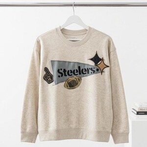 May include: A beige crew neck sweatshirt with a Steelers pennant design. The pennant features the word "Steelers", a football, and star-shaped embellishments. Additional patches include a foam finger. The sweatshirt is hanging on a white hanger.