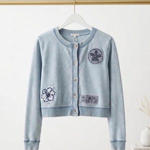 Puede incluir: Rebeca azul claro con botones y parches bordados. Cuello redondo, mangas largas y dobladillo corto. Parches con un diseño de flor y estrella de mar. La rebeca está colgada de una barra dorada.