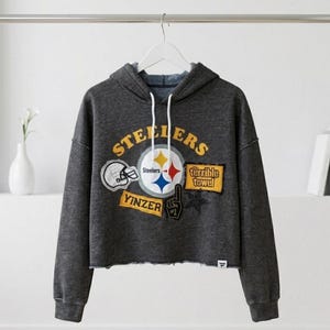 May include: Dark gray cropped hoodie with a drawstring hood. The front features multiple patches with the word "Steelers", a football helmet, the team logo, and the words "Terrible Towel" and "Yinzer".
