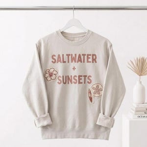 May include: A beige sweatshirt with the words "SALTWATER + SUNSETS" in a reddish-brown font. The sweatshirt features a floral patch and a seashell patch. The sleeves are cuffed at the wrists. The sweatshirt is hanging on a white hanger.