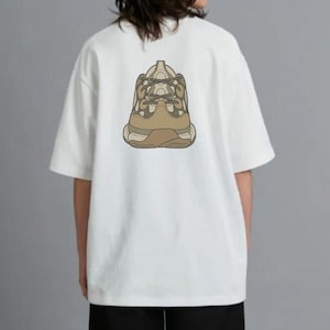 May include: White t-shirt with a graphic of a beige and brown sneaker on the back. The short-sleeved shirt is made of a soft material. The design features a detailed illustration of a chunky sneaker.