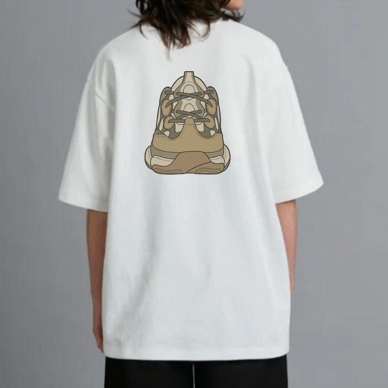 May include: White t-shirt with a graphic of a beige and brown sneaker on the back. The short-sleeved shirt is made of a soft material. The design features a detailed illustration of a chunky sneaker.