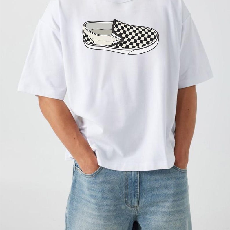 May include: White t-shirt featuring a black and white checkered slip-on shoe graphic. The graphic is a detailed illustration of the shoe's design. The shirt is paired with light blue denim jeans.