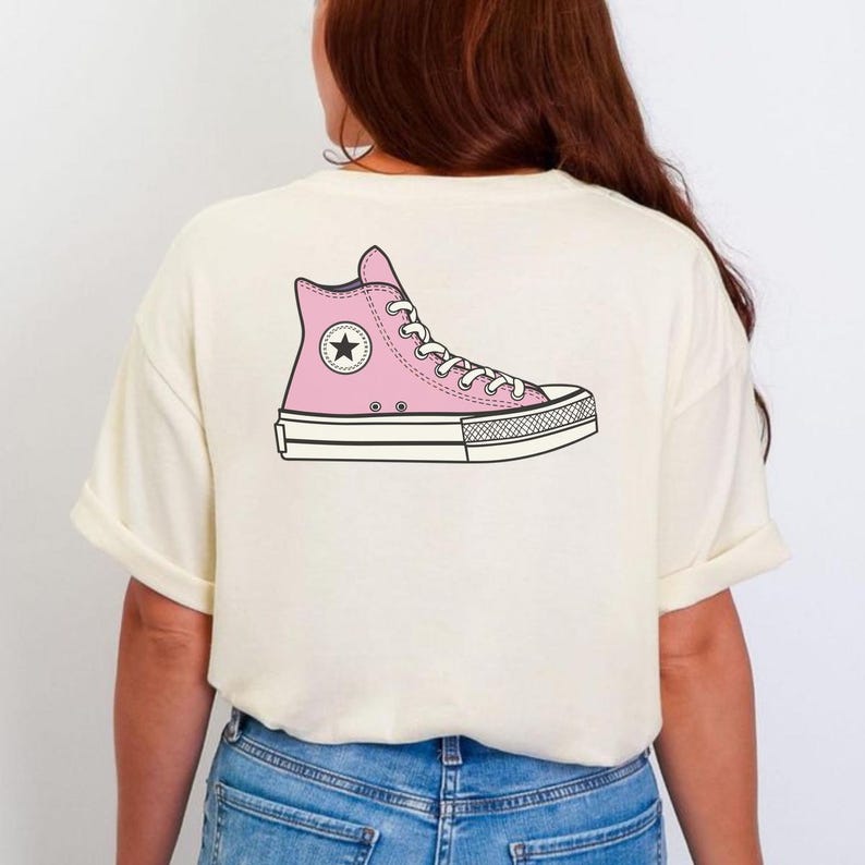 May include: Cream-colored t-shirt featuring a pink high-top sneaker graphic on the back. The sneaker design includes white laces, a black star emblem, and a black and white sole. The shirt has short sleeves and a relaxed fit.