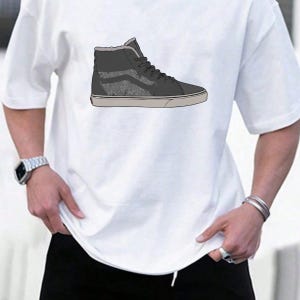 May include: White t-shirt featuring a high-top sneaker design. The sneaker is primarily dark gray with a textured pattern on the side and a light gray sole. The shoe has black laces and a light gray interior.