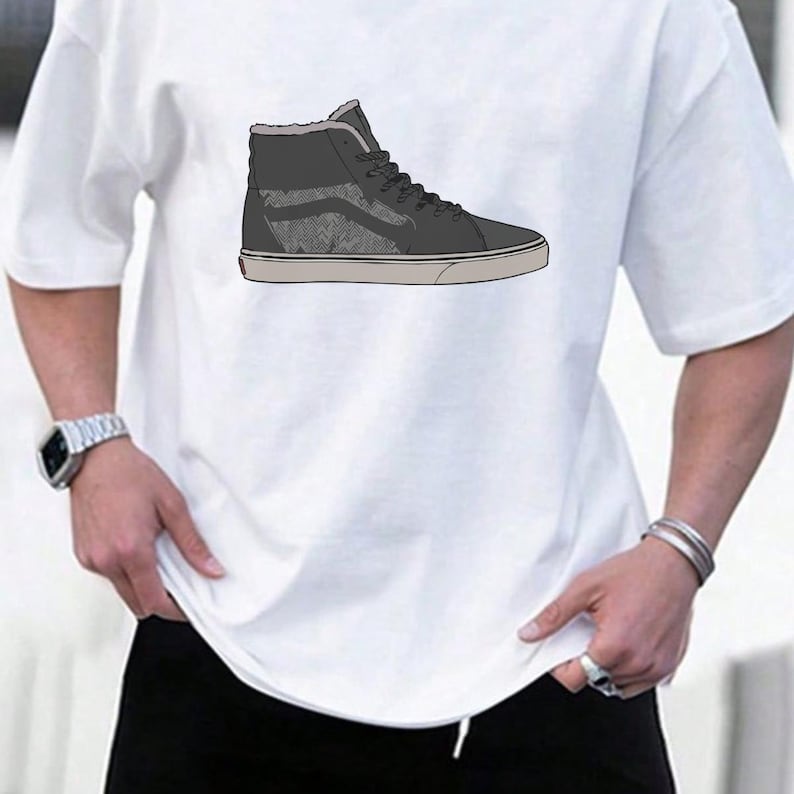 May include: White t-shirt featuring a high-top sneaker design. The sneaker is primarily dark gray with a textured pattern on the side and a light gray sole. The shoe has black laces and a light gray interior.