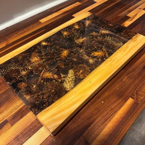 May include: A rectangular serving board featuring a central panel of dark, translucent resin embedded with numerous brown pinecones of varying sizes and textures, framed by two smooth, light-coloured wooden edges with a visible grain pattern.