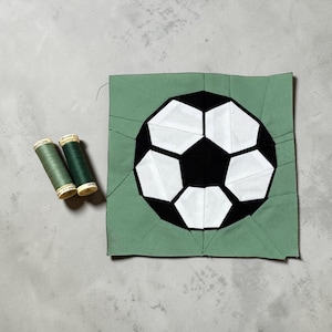 Puede incluir: Un parche de balón de fútbol hecho a mano con pentágonos negros y blancos, sobre un cuadrado verde. Dos carretes de hilo verde están junto al parche. El parche es probablemente para un proyecto textil.