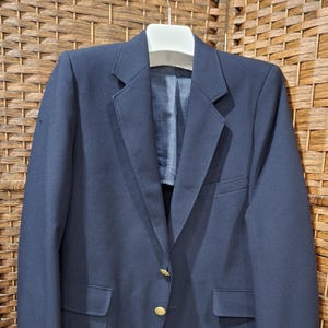 May include: A navy blue blazer with gold-colored buttons and two front pockets. The jacket is hanging on a white hanger against a woven background. The blazer has a classic, tailored design.