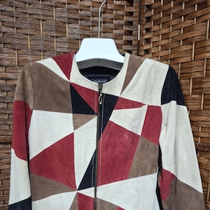 May include: A long-sleeve jacket featuring a geometric patchwork design in shades of cream, deep red, brown, and black. The jacket has a round neckline, a full-length front zipper closure with a metallic pull, and is constructed from suede panels.
