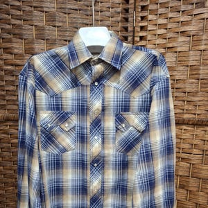 Puede incluir: Una camisa de cuadros de manga larga en tonos azul, beige y blanco. La camisa tiene dos bolsillos en el pecho con solapas puntiagudas y cierres de botón a presión de perlas. El cuello y los puños son visibles.