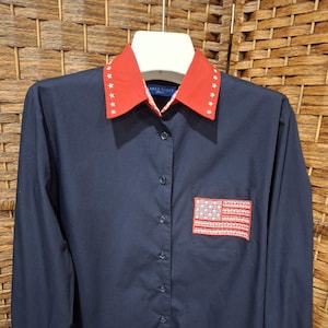 May include: Navy blue button-down shirt with a red collar adorned with white stars. The shirt features a small American flag patch on the chest. The long-sleeved shirt is made of a smooth fabric and is hanging on a white hanger.