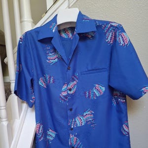 Può includere: Una camicia blu a maniche corte con un motivo di ananas colorati. La camicia ha un colletto classico, una tasca sul petto e bottoni neri. Gli ananas sono nelle tonalità del rosa, del blu e del verde acqua.