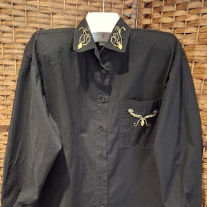 May include: Black button-down shirt with long sleeves and a white collar. The collar and pocket feature gold embroidery. The shirt is hanging on a white hanger against a woven background. The shirt has a single pocket on the left side.