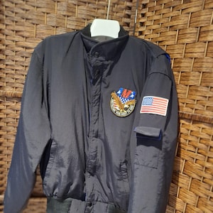 May include: A navy blue bomber jacket with a black collar and cuffs. The jacket features a patch with an eagle, the American flag, and the text "NIGHT RIDERS". It has a button-up front and a small pocket on the sleeve.