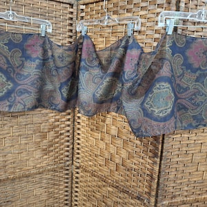 May include: Three paisley patterned scarves in shades of navy blue, brown, and pink, hanging on clear plastic hangers. The scarves have a repeating floral design. The background is a woven wicker screen.