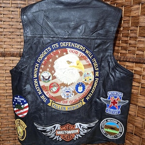 Könnte beinhalten: Schwarze Lederweste mit mehreren Aufnähern. Aufnäher sind ein Weißkopfseeadler, die Worte "The Nation Which Forgets Its Defenders Will Itself Be Forgotten" und ein Harley-Davidson-Logo. Weitere Aufnäher zeigen militärische Embleme.