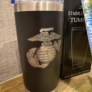 May include: Black stainless steel tumbler with a silver rim and lid. The tumbler features an engraved emblem of an eagle over a globe with the words "SEMPER FIDELIS". The box next to it reads "STAINLESS TUMBLER" and "Double Wall For Cold & Hot Drinks".
