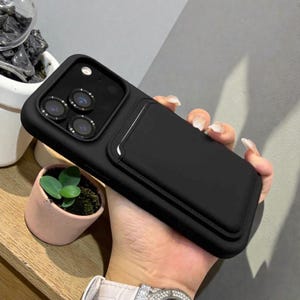 Può includere: Una custodia per telefono nera con un portacarte integrato. La custodia ha una finitura opaca e liscia e un ritaglio per la fotocamera per più obiettivi. Il portacarte è rettangolare e sembra essere realizzato con un materiale simile. La custodia è tenuta da una persona.