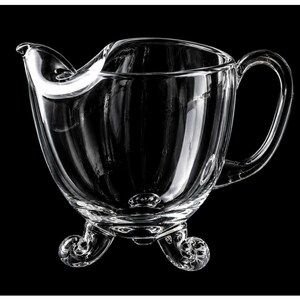 May include: A clear glass creamer with a curved spout and a handle. The creamer sits on three decorative feet. The glass is transparent, allowing light to pass through, and is set against a black background. The creamer is designed for serving liquids.