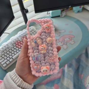 May include: A pink phone case with a decorative design. The case has a textured, layered pattern with small, colourful embellishments, including cartoon characters, butterflies, and hearts. The case is designed to fit a smartphone.