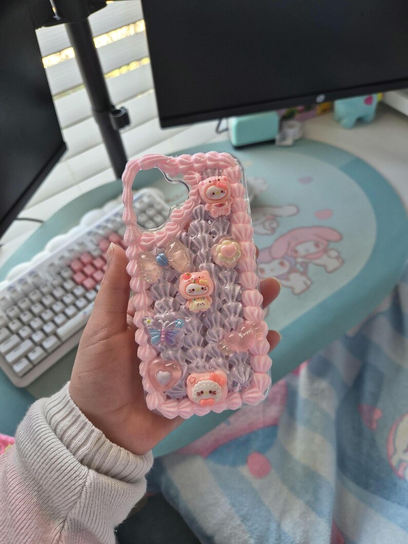 May include: A pink phone case with a decorative design. The case has a textured, layered pattern with small, colourful embellishments, including cartoon characters, butterflies, and hearts. The case is designed to fit a smartphone.