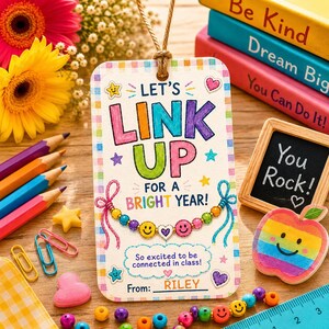 Puede incluir: Una etiqueta de regalo rectangular con borde a cuadros que presenta el texto 'LET'S LINK UP FOR A BRIGHT YEAR!' y 'So excited to be connected in class! From: RILEY'. La etiqueta muestra un gráfico de pulsera de cuentas coloridas con caras sonrientes.