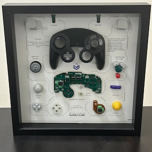 May include: Framed Nintendo GameCube controller parts display. Includes a black controller, circuit board, buttons, and other components. The text "NINTENDO GAMECUBE" is visible. The frame is black.
