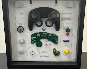 May include: Framed Nintendo GameCube controller parts display. Includes a black controller, circuit board, buttons, and other components. The text "NINTENDO GAMECUBE" is visible. The frame is black.
