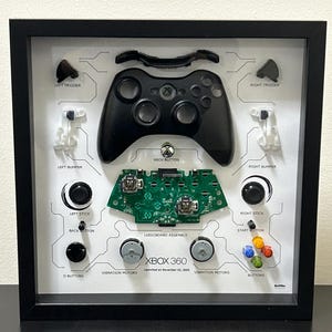 May include: Framed Xbox 360 controller parts display. The black controller is centered, with labeled components like triggers, bumpers, and sticks. The green logic board and vibration motors are also visible. The frame is black.