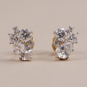 May include: A pair of stud earrings featuring a cluster of clear, faceted stones in various shapes, including round, pear, and square cuts, set in a metallic base. The stones are arranged in an asymmetrical pattern, creating a sparkling, multi-faceted appearance on each earring.