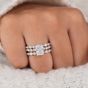 May include: A three-piece silver-toned ring set featuring a central rectangular radiant-cut stone flanked by two matching bands. Each band is adorned with a series of small, marquise-cut stones arranged in a continuous row, creating a cohesive look.