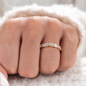 May include: A gold band ring featuring a series of clear, rectangular-cut stones set in a bezel style. The stones are arranged horizontally along the top half of the band, creating a continuous, linear design with a polished metallic finish.