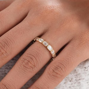 May include: A gold band ring featuring a series of clear, rectangular-cut stones set in individual bezels. The stones are arranged in a continuous row around the band, creating a uniform, linked appearance with a polished metallic finish.