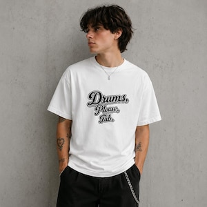 May include: A white t-shirt featuring the text "Drums, Please, Fab" in a black, decorative font. The t-shirt has short sleeves and a loose fit. The image shows the t-shirt on a person.