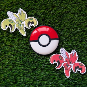 May include: A red, white, and black Poké Ball sits between two character stickers. One sticker is a green and yellow insect-like creature, and the other is a red insect-like creature. The items are on a green, textured surface.