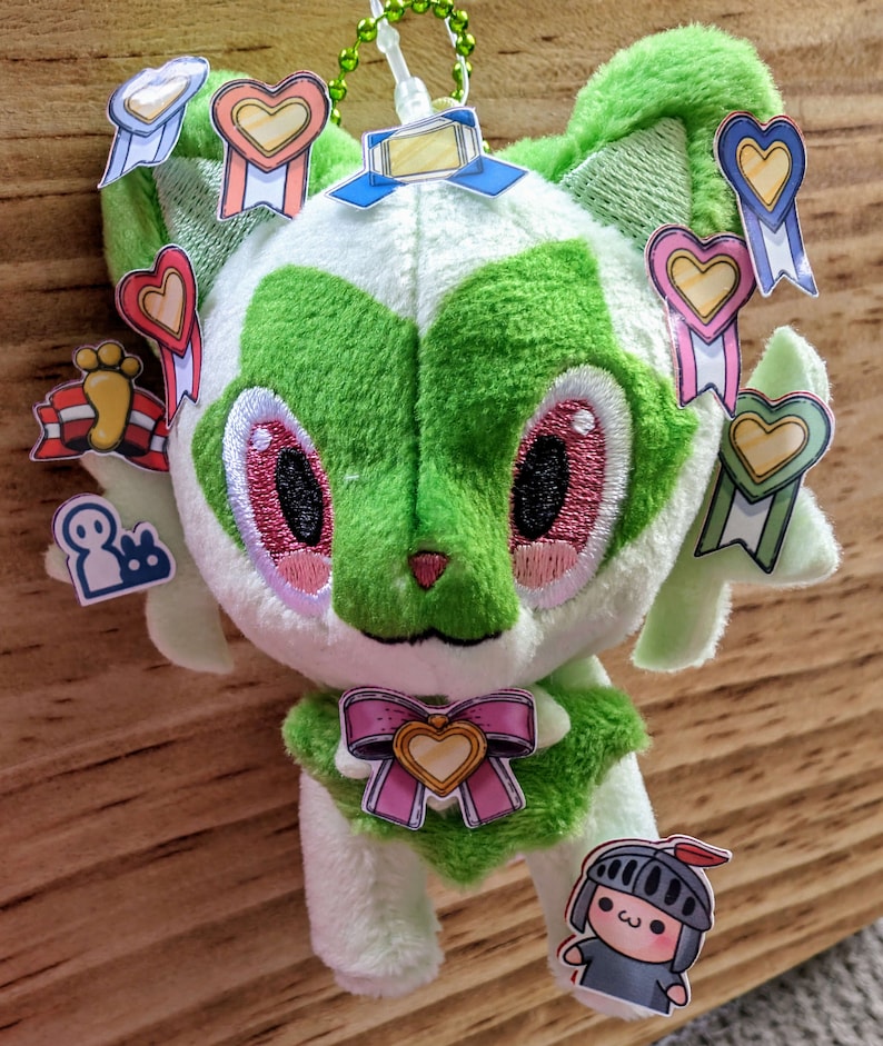 Pode incluir: Um brinquedo de pel&uacute;cia verde e branco com olhos bordados e um la&ccedil;o rosa. O brinquedo tem v&aacute;rios adesivos coloridos anexados a ele, incluindo cora&ccedil;&otilde;es, estrelas e um cavaleiro.