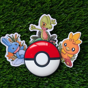 May include: A collection of colorful stickers featuring cartoon characters and a red, white, and black Poké Ball. The stickers are placed on a green, grassy surface. The characters include a blue and white creature, a green lizard, and an orange bird.