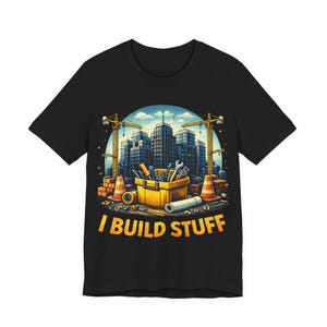 Könnte beinhalten: Schwarzes T-Shirt mit einer farbenfrohen Grafik einer Baustelle. Das Design zeigt eine gelbe Werkzeugkiste voller Werkzeuge, Leitkegel und eine Stadtlandschaft mit Kränen. Der Text "I BUILD STUFF" ist in Gelb gedruckt.