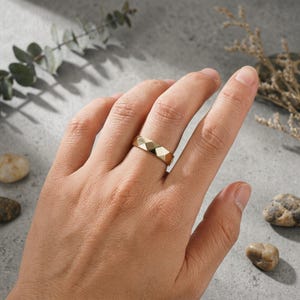 May include: A gold-colored ring with a geometric, faceted design. The ring is shown on a hand, with a light gray background and decorative elements. The ring's design features multiple flat surfaces, creating a textured appearance.