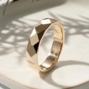 May include: A gold ring with a geometric, faceted design. The ring has a polished finish and a band with multiple flat surfaces. The ring is set on a white surface, with shadows in the background.