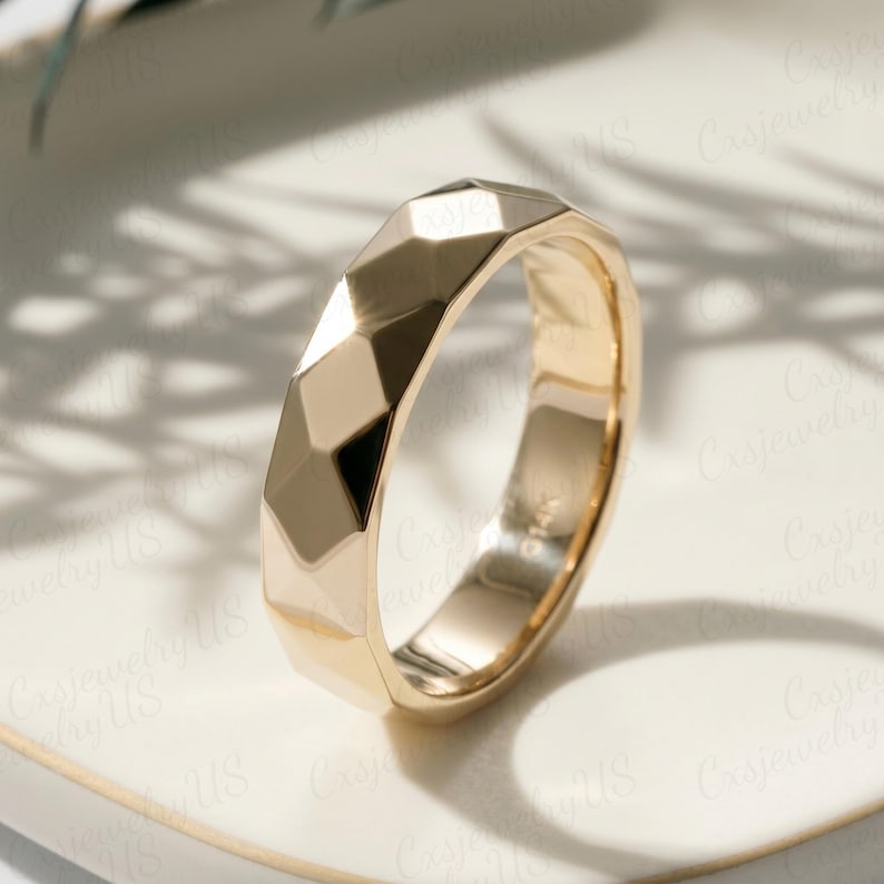 May include: A gold ring with a geometric, faceted design. The ring has a polished finish and a band with multiple flat surfaces. The ring is set on a white surface, with shadows in the background.