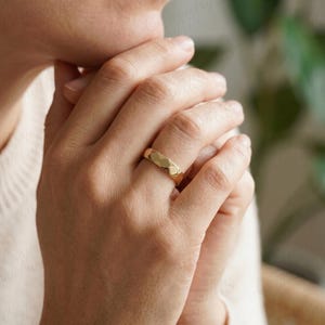 May include: A gold-colored ring with a geometric, faceted design. The ring is worn on a finger, showcasing its textured surface and reflective qualities. The ring's design features a series of flat, angled surfaces that catch the light.