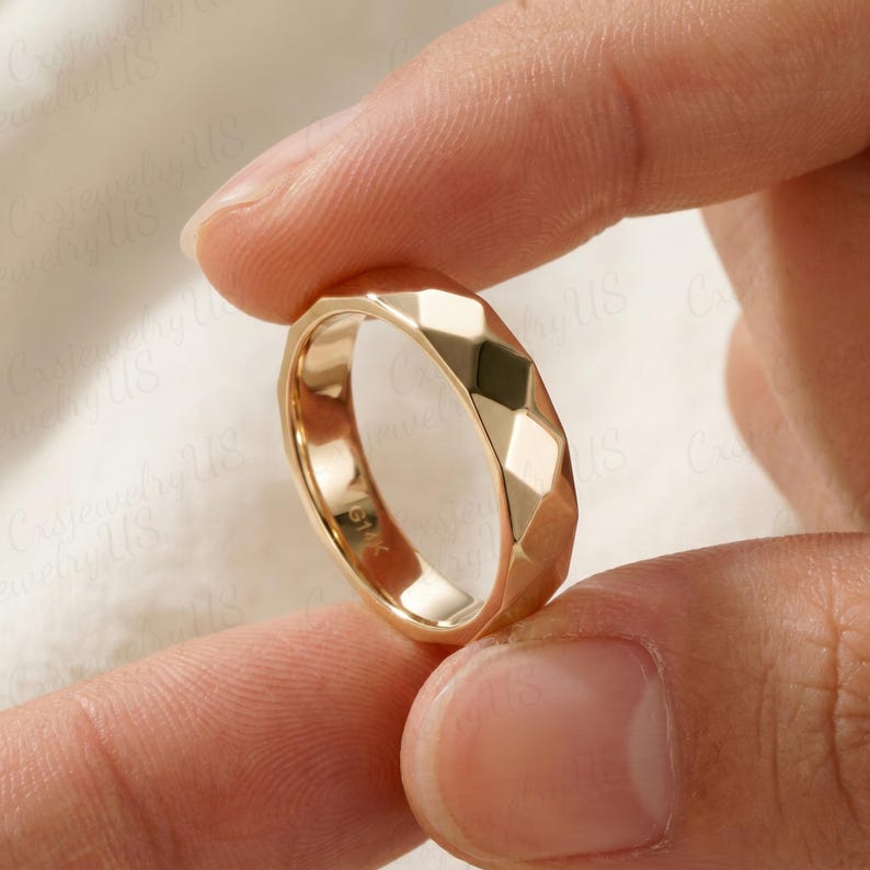 May include: A gold ring with a geometric, faceted design. The ring is held between two fingers, showcasing its polished surface and intricate pattern. The ring is marked with "G14K".