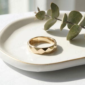 May include: A gold-colored ring with a geometric design, featuring a series of angled facets around the band. The ring is placed on a white, oval-shaped dish, with a sprig of greenery in the background. The ring is marked with "G14K".
