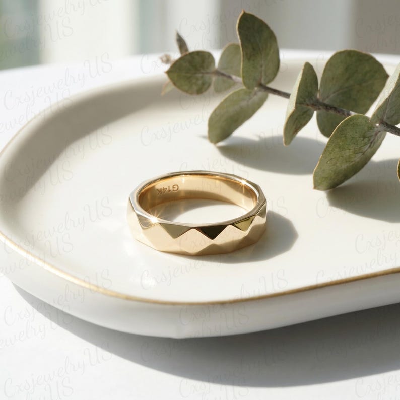May include: A gold-colored ring with a geometric design, featuring a series of angled facets around the band. The ring is placed on a white, oval-shaped dish, with a sprig of greenery in the background. The ring is marked with "G14K".