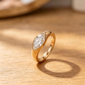 Op de afbeelding: Een gouden ring met een marquise-geslepen diamant. De diamant is gezet in een bezel-zetting en de band is glad en gepolijst. De ring wordt gepresenteerd op een houten oppervlak, met natuurlijke schaduwen.