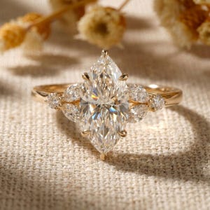 May include: A gold engagement ring with a large, marquise-cut diamond as the centerpiece. The ring features smaller, pear-shaped diamonds on either side of the main stone, set on a delicate band. The ring is displayed on a neutral-colored fabric.