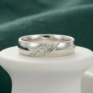 Puede incluir: Un anillo plateado con una línea diagonal de pequeñas piedras preciosas transparentes. El anillo está sobre una superficie blanca, con un fondo verde oscuro. La banda es lisa y pulida.