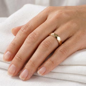 May include: A gold ring with a geometric, faceted design. The ring is a warm gold color and has a series of flat, angled surfaces that catch the light. The ring is on a finger, resting on a white cloth.